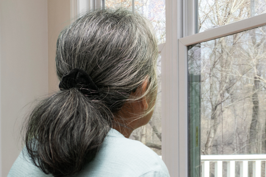 older woman looks out of a window with trees visible.